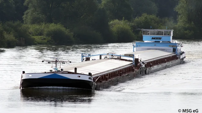 Ein Frachtschiff mit flachem Aufbau fährt auf einem Fluss. Die Oberfläche des Schiffes ist größtenteils mit hellen Abdeckungen versehen. Im Hintergrund sind dicht bewachsene grüne Uferbereiche zu sehen. Das Wasser reflektiert das Tageslicht.