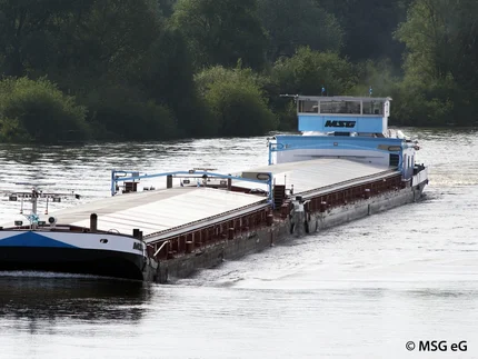 Ein Frachtschiff mit flachem Aufbau fährt auf einem Fluss. Die Oberfläche des Schiffes ist größtenteils mit hellen Abdeckungen versehen. Im Hintergrund sind dicht bewachsene grüne Uferbereiche zu sehen. Das Wasser reflektiert das Tageslicht.