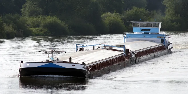 Ein Frachtschiff mit flachem Aufbau fährt auf einem Fluss. Die Oberfläche des Schiffes ist größtenteils mit hellen Abdeckungen versehen. Im Hintergrund sind dicht bewachsene grüne Uferbereiche zu sehen. Das Wasser reflektiert das Tageslicht.