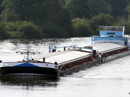 Ein Frachtschiff mit flachem Aufbau fährt auf einem Fluss. Die Oberfläche des Schiffes ist größtenteils mit hellen Abdeckungen versehen. Im Hintergrund sind dicht bewachsene grüne Uferbereiche zu sehen. Das Wasser reflektiert das Tageslicht.